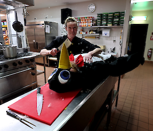 Lady dressed in chef blacks and holding cleaver stands over man in penguin costume lying on kitchen bench