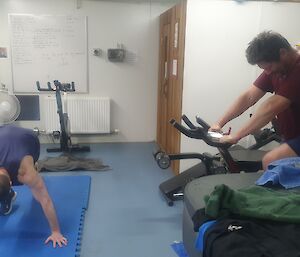 A man riding a bike and another does burpees on a mat on the floor