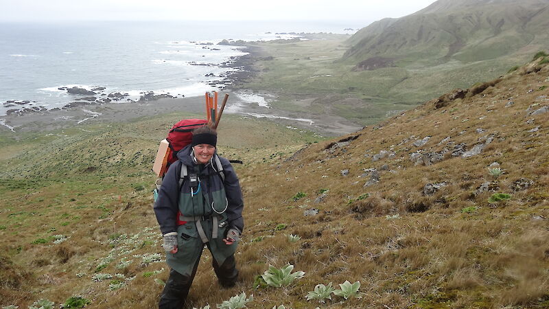 A woman in outdoor survival gear smiles at the camera. She is standing on a grassy slop with cliffs, ocean and rocky beach behind her