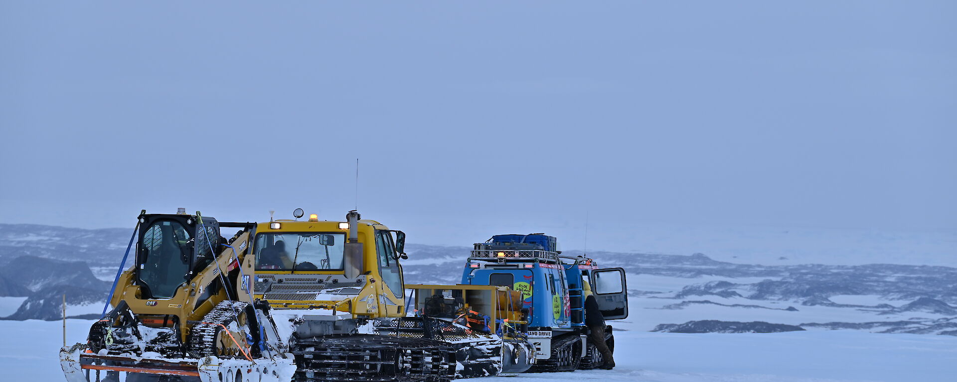 A blue Hägglunds and a snow groomer towing a sled on the sea ice with hills in the background