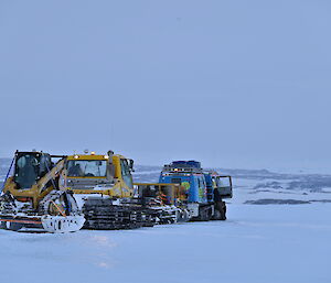 A blue Hägglunds and a snow groomer towing a sled on the sea ice with hills in the background