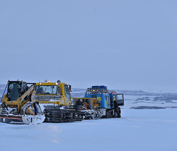 A blue Hägglunds and a snow groomer towing a sled on the sea ice with hills in the background