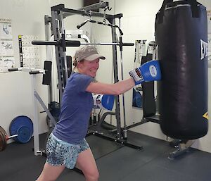 Woman in weight gym with boxing gloves on, smiles at camera as punches heavy punching bag hanging from ceiling