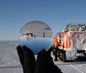 Crystal ball held up by hand in black glove show reversed image of Hagglunds and two people on sea ice