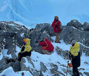 Four people clamber over rocks to get good view of emperor penguins in the distance with glacier cliffs rising up behind