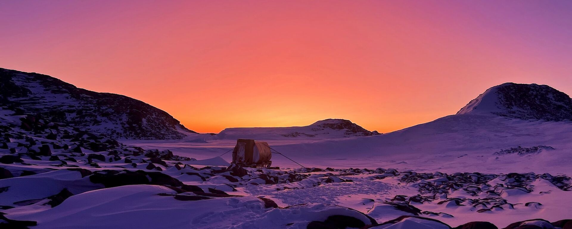 Ice covered ground with small hut in center of picture, hills in the background. The light is intense pinks, purples and oranges as sun rises
