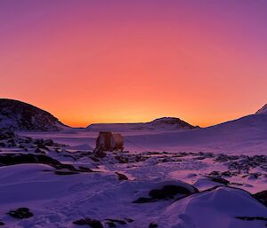 Ice covered ground with small hut in center of picture, hills in the background. The light is intense pinks, purples and oranges as sun rises
