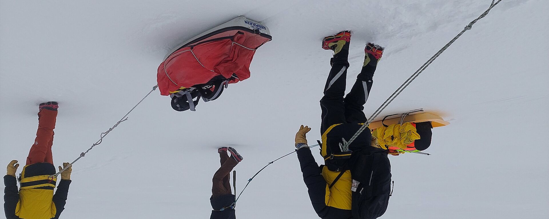 Three people in thick winter clothing walking over a snowy plain. They are pulling small sleds loaded with gear behind them, by ropes attached to their waists