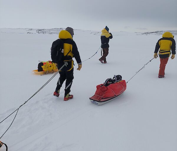 Three people in thick winter clothing walking over a snowy plain. They are pulling small sleds loaded with gear behind them, by ropes attached to their waists