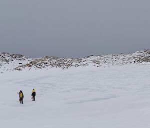 A gentle, snowy downslope to a frozen lake with low, rocky, snow-dusted hillocks by its far shore. Four people can be seen walking towards the lake, three in the distance approaching the lake's shore, and one far behind them