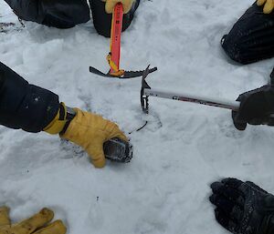 A close-up picture of a small handheld device lying on snowy ground. A person (out of frame) wearing thick, yellow gloves is picking up the device. Ice axes, a shovel handle and other gloved hands can be seen around the edge of the picture, suggesting a group of people kneeling around the spot where the device is found.