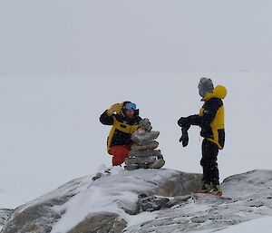 Two people in thick winter clothing, crouching and standing on top of a snow-covered rock. One person is building a small cairn out of a number of flat stones, while the other person looks on
