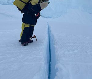 A person in thick winter clothing with a pack on her back is standing on thick sea ice and looking down into a tide crack. The tide crack looks like a narrow but deep, bluish fissure in solid white ground