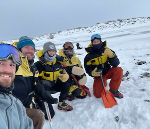 A selfie-angled photo of five people in thick winter clothing, kneeling in the snow beside a small handheld device they have just discovered on the ground. The people are carrying ice axes and shovels and smiling at the camera
