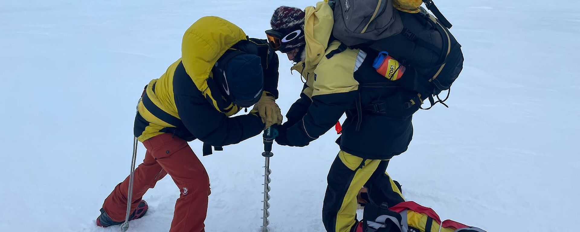 Two people standing on a wide, white stretch of sea ice. One person is operating a powered hand drill with a long flight attached, to drill down into the ice. The other person is pressing the drill apparatus down into the ice with both hands