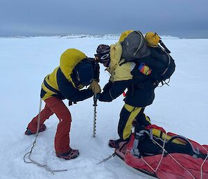 Two people standing on a wide, white stretch of sea ice. One person is operating a powered hand drill with a long flight attached, to drill down into the ice. The other person is pressing the drill apparatus down into the ice with both hands