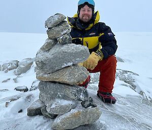 A person in thick winter clothing, smiling at the camera as he kneels beside a small stone cairn he has constructed