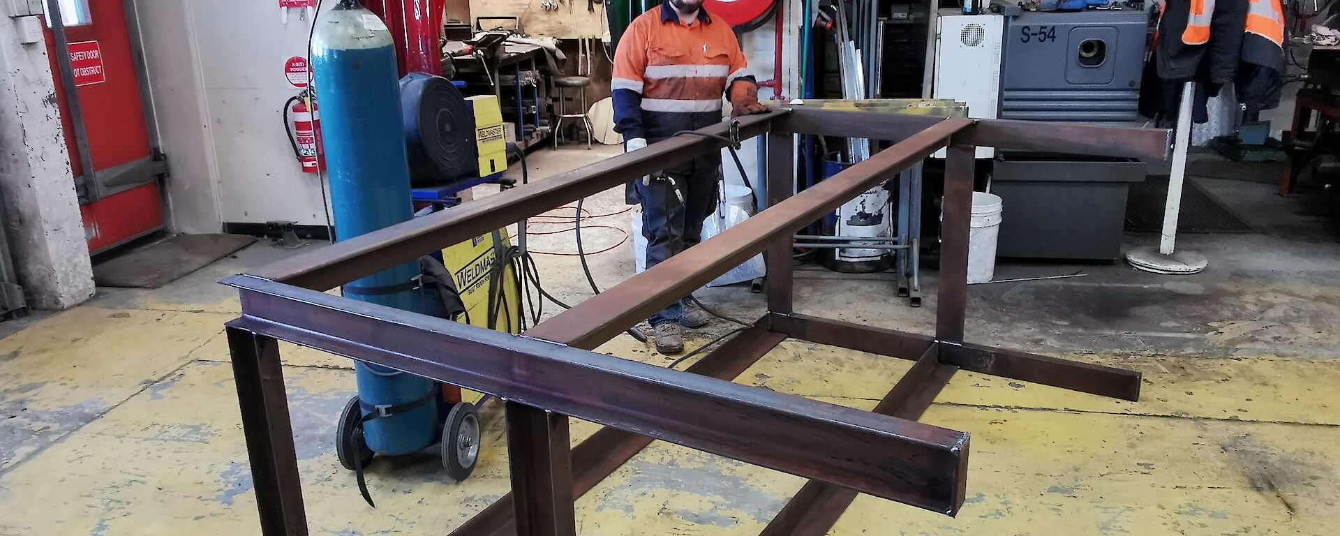 A man in hi-vis standing behind a steel structure with some welding equipment in a workshop