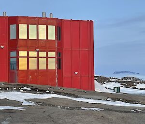 Rocky foreground, large red square building with multiple windows in centre, mountain range in background.  Patches of snow can be seen on the ground.