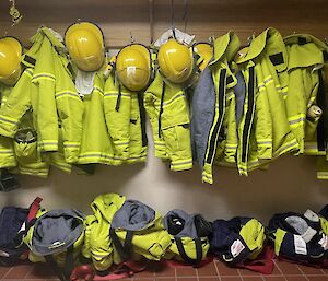 Fire turn-out gear (yellow jackets and helmets) hanging on hooks and below row of boots with yellow fire pants in position ready to be stepped into