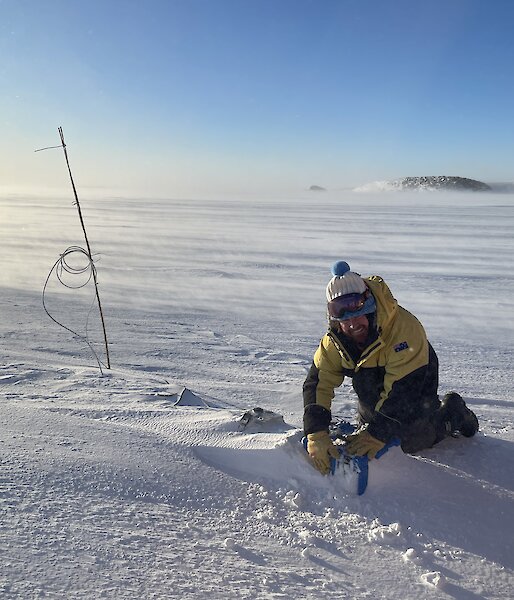 Man in winter gear in a snow covered landscape kneels over a mound in sea-ice checking instruments