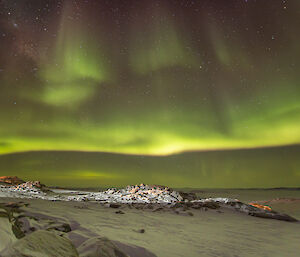 The night sky over a snowy, rocky ground sloping down to an icebound ocean. The scene appears to be lit brightly by an aurora, broad, green swathes of light fading upwards into dim red