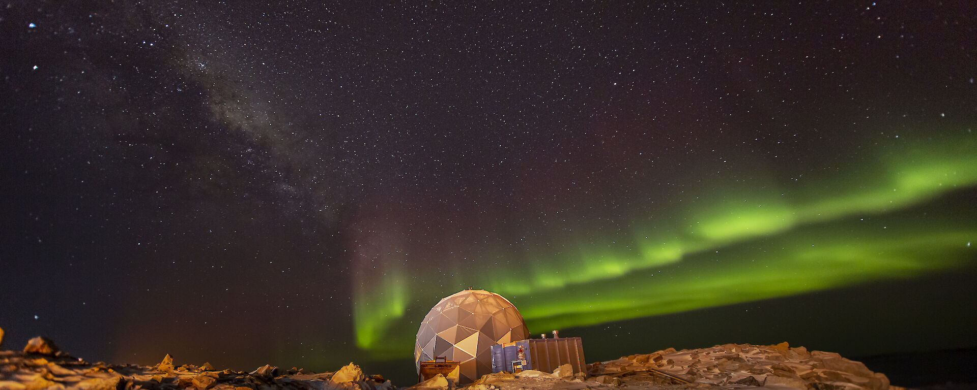 A polyhedral structure on rocky, snowy ground, beneath a night sky lit up with an aurora, presently showing as two parallel bands of bright green light