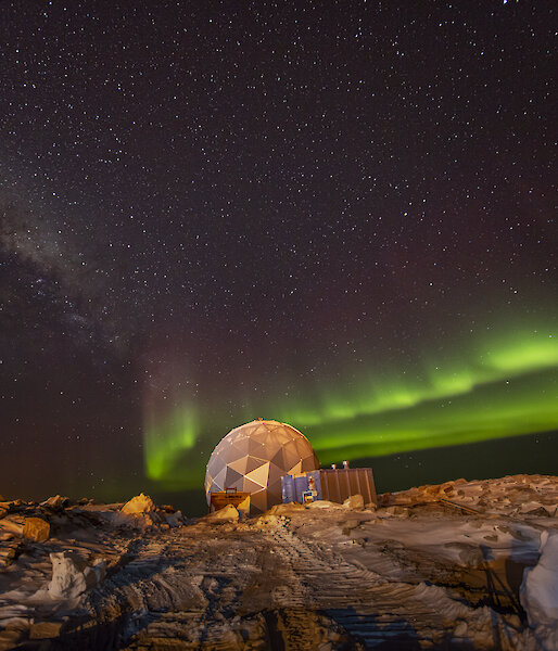 A polyhedral structure on rocky, snowy ground, beneath a night sky lit up with an aurora, presently showing as two parallel bands of bright green light