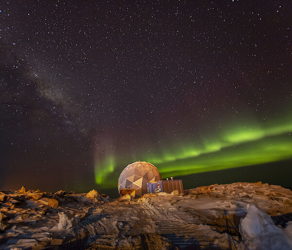 A polyhedral structure on rocky, snowy ground, beneath a night sky lit up with an aurora, presently showing as two parallel bands of bright green light