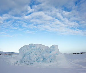 A small iceberg locked fast into white sea ice. The iceberg is riddled with holes varying in depth and size, making a cyan-blue-shadowed texture on its white surfaces. The blue sky above is partly covered with a patchy layer of cloud