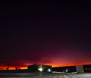 Early dawn scene at an Antarctic station. Above a large green shed, the sky is mostly dark, but the first light reflected off high clouds is in dark tones of red