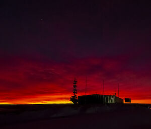 Early dawn scene at an Antarctic station. Above a large green shed, the first light reflected off a thick layer of clouds is deep scarlet