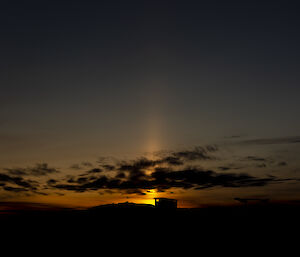 A dusk scene. The sun has just set, and appears to project a pillar of light vertically from the horizon. Thick, wispy-edged tufts of high cloud float in front of the pillar