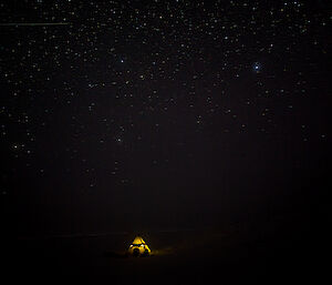 A night camping scene in almost complete darkness, except for small pyramidal tent lit from within and the stars in the sky