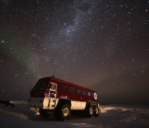 A red bus on extra large tyres parked on snow-covered ground. The night sky above is thick with stars. A few streaks of aurora show like faint rainbows