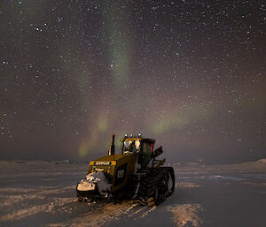 A yellow Caterpillar tractor parked on snowy ground. The night sky above is thick with stars. A few streaks of aurora show in faded greens and reds