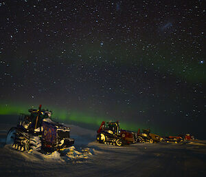 A row of brightly-painted tractors parked on snowy ground, beneath the night sky. A couple of horizontal bands of green light in the sky show aurora activity