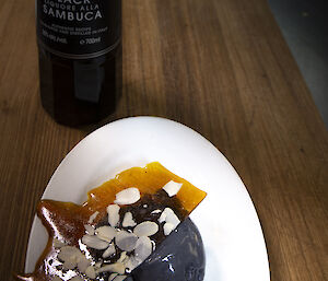 An artistically-plated dessert of grey ice cream and dried blueberries, topped with a thin sheet of toffee coated with almond flakes. Next to the plate is a black bottle with a label reading, "Galliano Black Liquore Alla Sambuca"