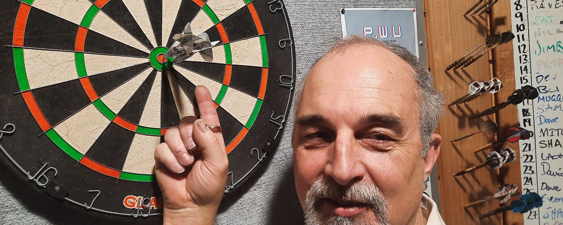 A close-up of a dartboard with two darts in the bullseye. The man who apparently threw those darts stands close by, pointing at them