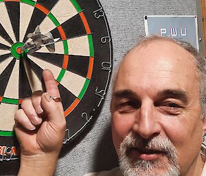 A close-up of a dartboard with two darts in the bullseye. The man who apparently threw those darts stands close by, pointing at them
