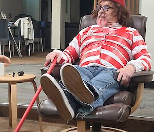 A man in a "Where's Wally" costume, comprising of a red-and-white-striped top, a red pointed beanie, thick-framed glasses and a red cane, reclining in an armchair