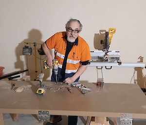 A man in a workshop wearing an orange work shirt, using a hand drill to drill into a panel of wood balanced on trestles