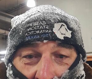 A selfie shot of a man in a black beanie and a face buff pulled down to reveal his eyes and nose. The beanie and buff are coated with clumps and flakes of ice, indicating that he has been outdoors in very cold weather