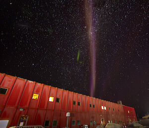 A long red rectangular building, photographed at night. From behind the building, a long, thin purplish plume of light appears to reach into the sky