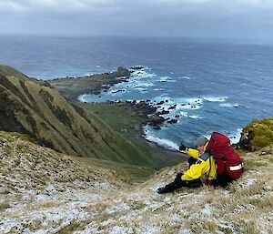 A man sits at the top of a steep slope heading down towards the sea