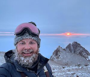 Man with ice covered beard and beanie, on top of rocky peak with ice in distance to horizon at sunset