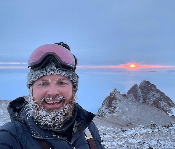 Man with ice covered beard and beanie, on top of rocky peak with ice in distance to horizon at sunset