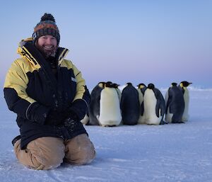 Man kneels in foreground, closely behind a group of emperor penguins poses for the camera
