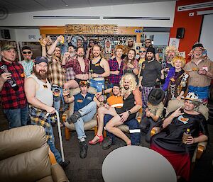 A group of about 25 people, many of them in checked flannel shirts, some wearing blonde, curly wigs, assembled close together in a lounge area for a group photo