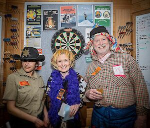 Three people in costume standing together before a dartboard. One wears a sheriff hat, another wears a purple feather boa, and the third wears a colourful cloth headdress. The last also has a piece of notepaper with a simple drawing of a cat's head and the word "meow" sticky-taped to his shirt